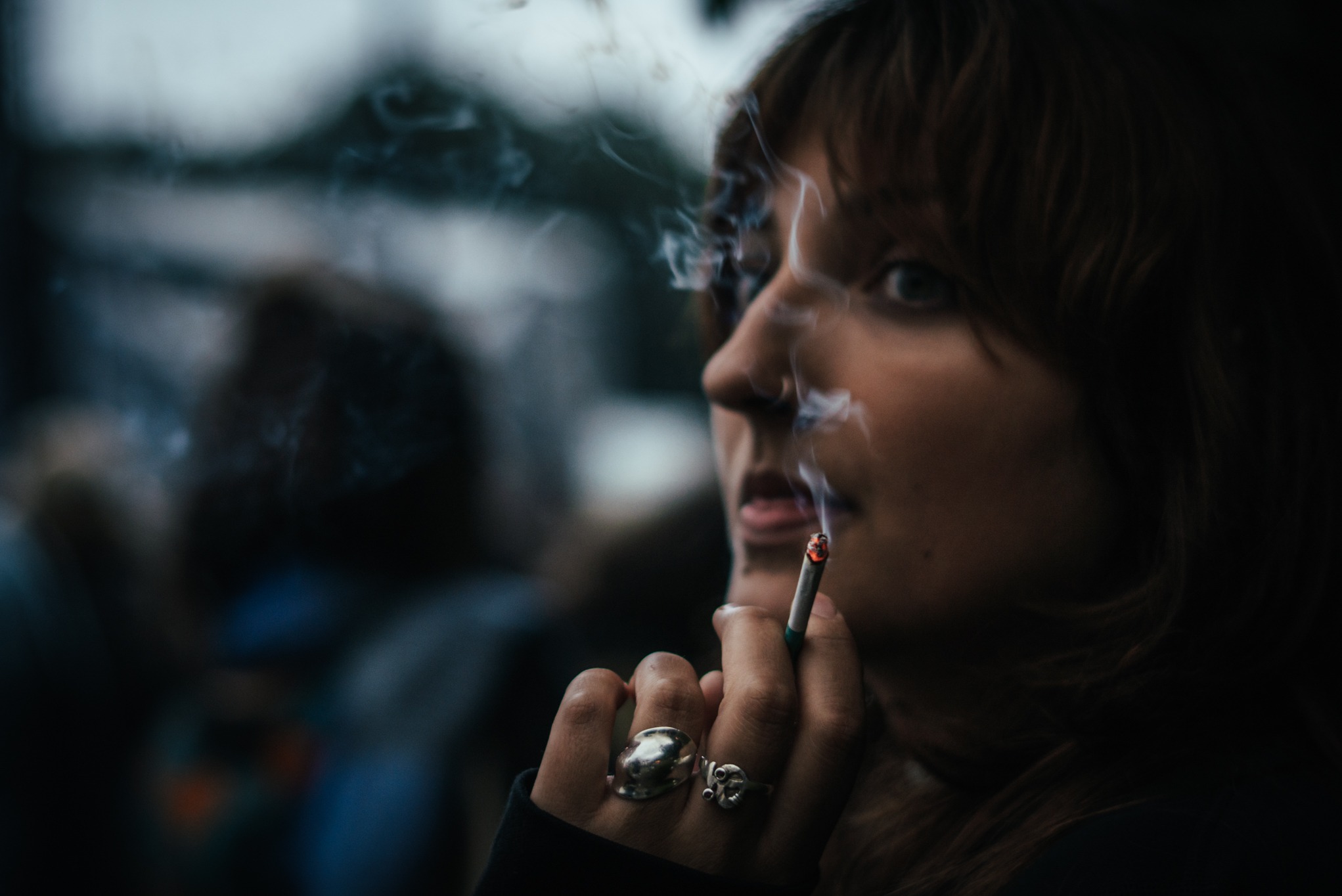 Portret kobiety z papierosem w ustach, dym unosi się w powietrzu. Widoczne srebrne pierścionki na palcach, ostre światło pada na profil twarzy.