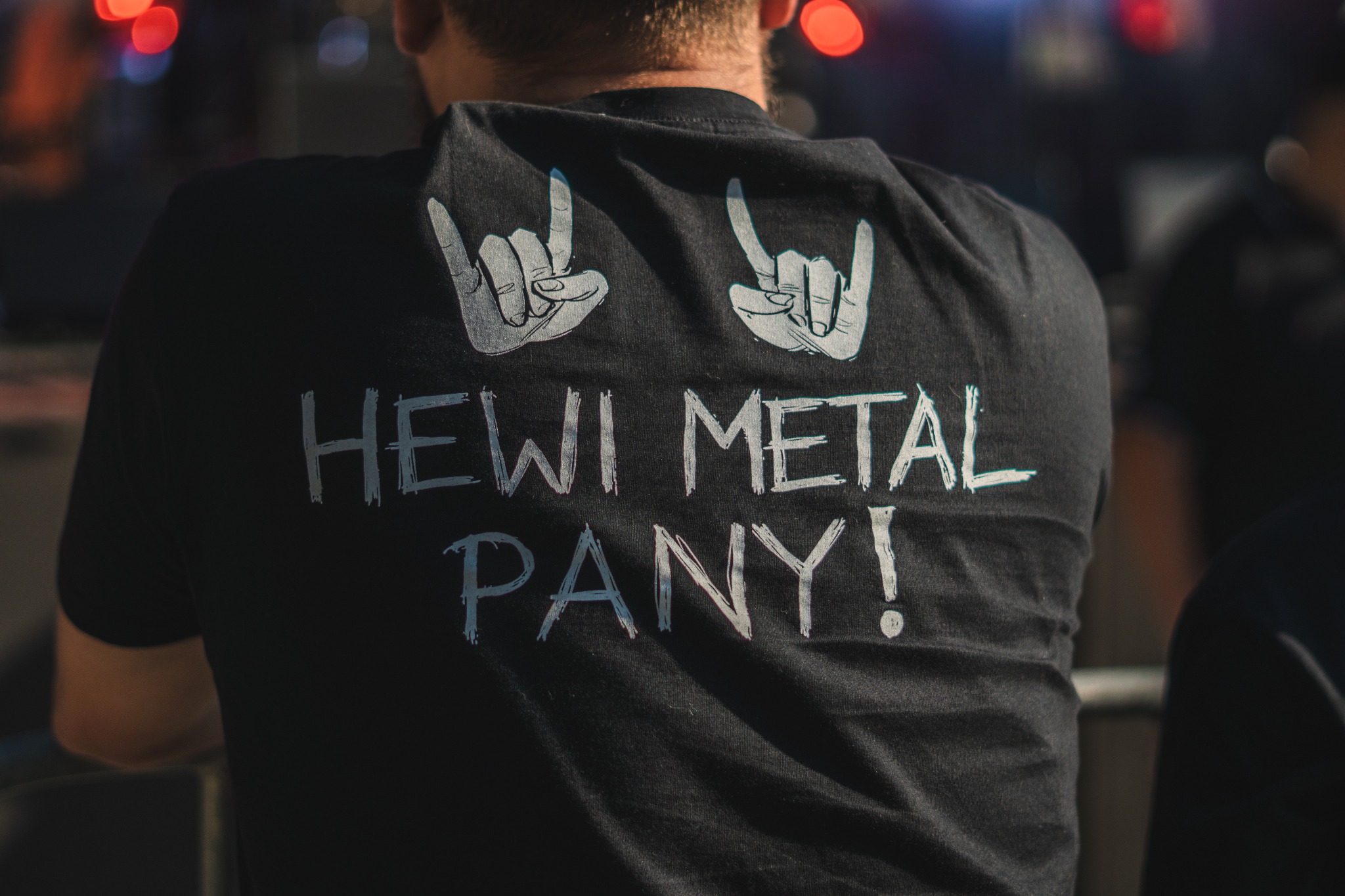 Plecy mężczyzny w czarnej koszulce z białym napisem 'HEWI METAL PANY!' i dwoma symbolami 'diabelskich rogów' na tle rozmytych świateł.
