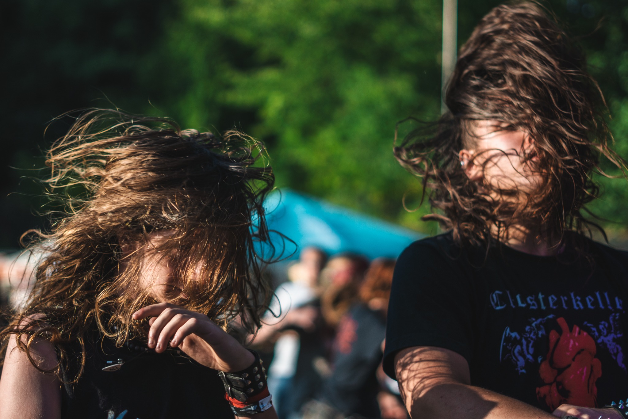 Dwie osoby z długimi włosami w ruchu na tle zieleni i rozmytych postaci, ujęte w dynamicznym kadrze podczas wydarzenia muzycznego.
