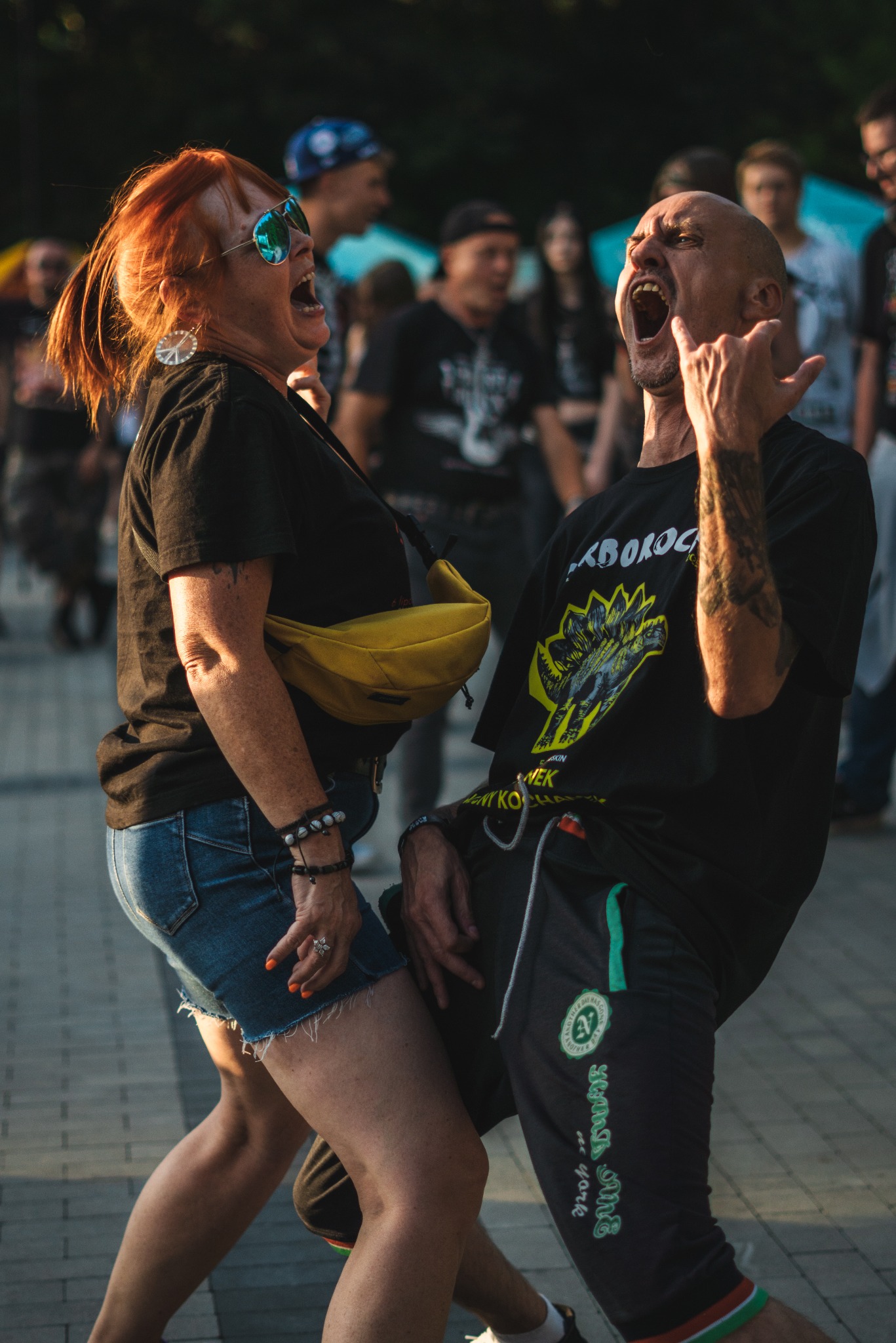 Ekspresyjne ujęcie pary na koncercie: kobieta w okularach i mężczyzna z tatuażem, oboje z otwartymi ustami w uniesieniu, energia sceny i tłumu. Ulica, światło słoneczne.