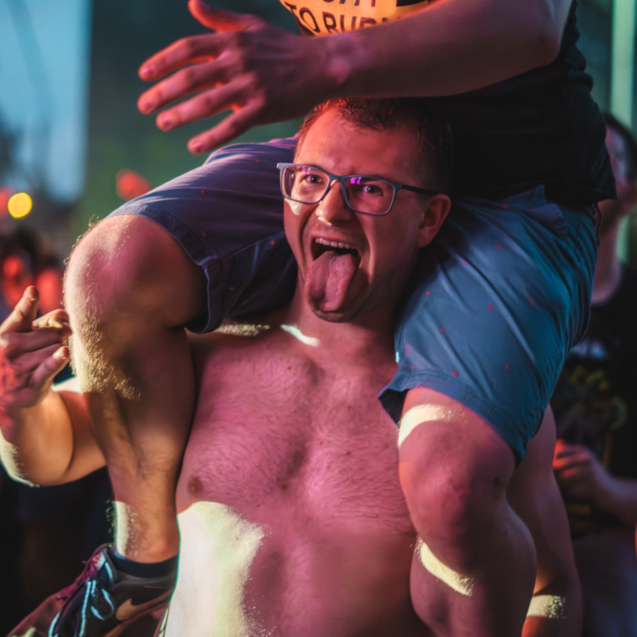 Rozbawiony mężczyzna w okularach, bez koszuli, niesiony na barana podczas imprezy. W tle widoczni inni uczestnicy i kolorowe światła.