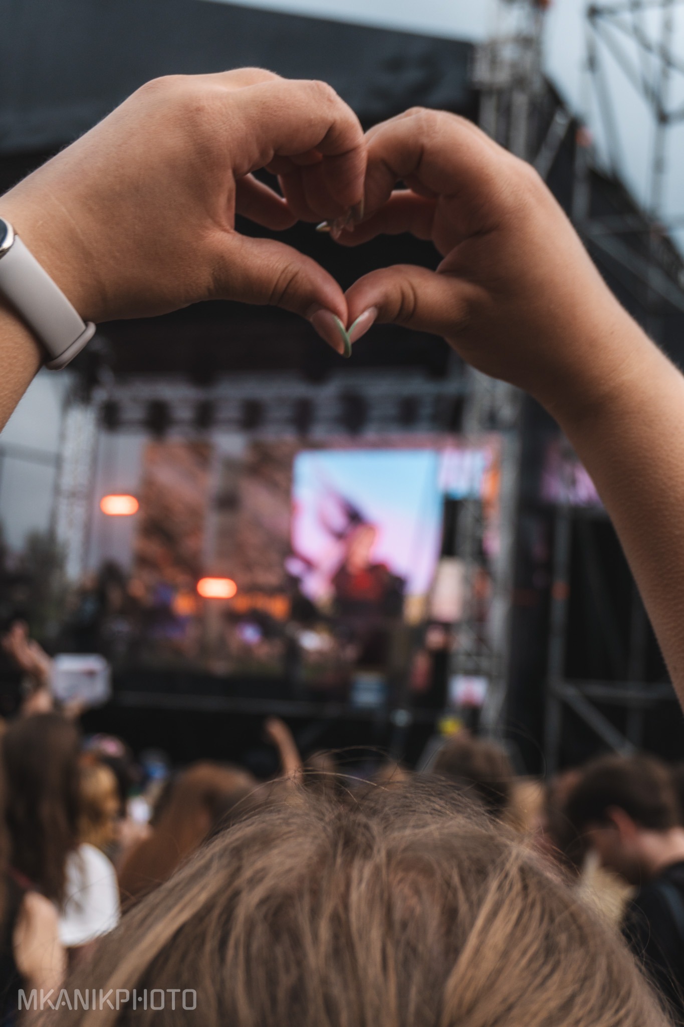 Ręce formujące serce na tle sceny koncertowej z rozmytą publicznością. Widoczny zegarek na nadgarstku i jasny manicure. Atmosfera wydarzenia.