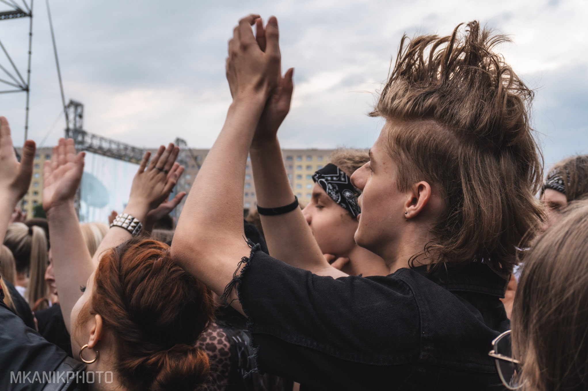 Tłum ludzi z podniesionymi rękami na koncercie. W centrum młody mężczyzna z irokezem, profil, ciemne ubranie. W tle scena i elementy konstrukcji scenicznej.