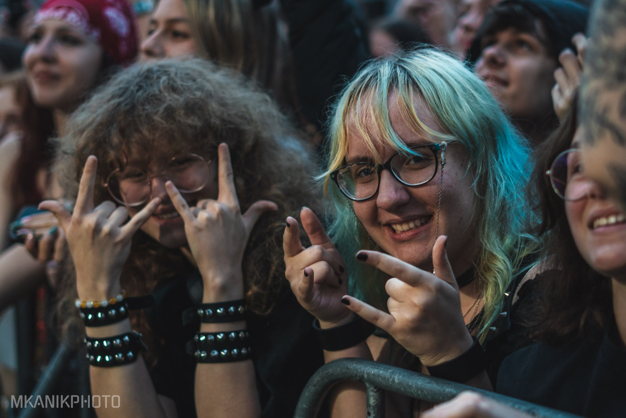 Rozradowane fanki z okularami i rockowymi gestami na koncercie, w tłumie, z widocznym znakiem wodnym fotografa.