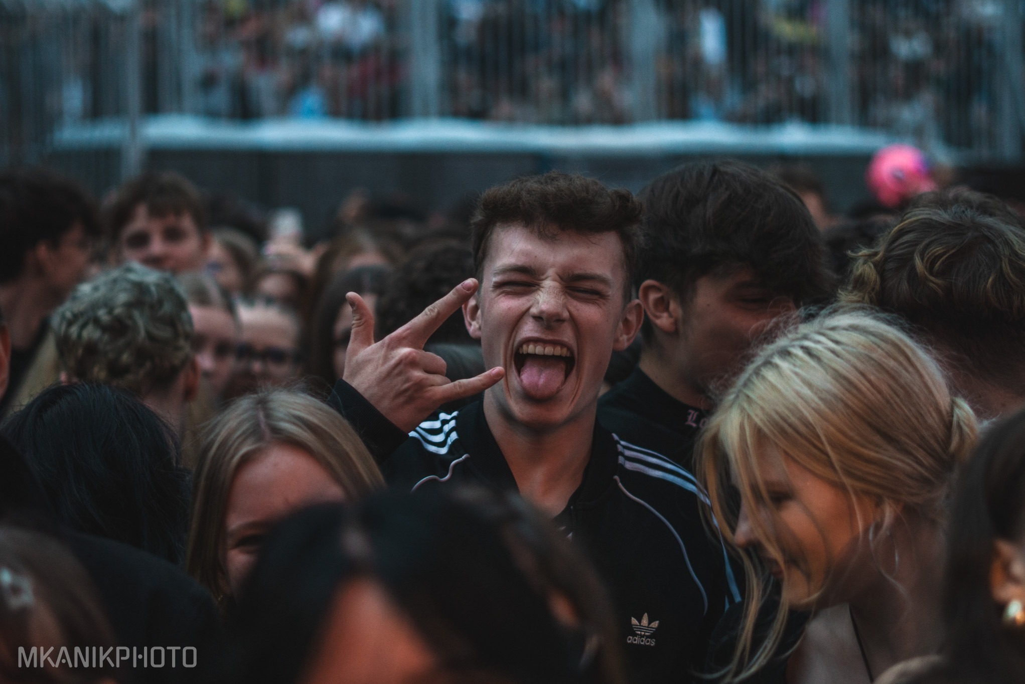 Młody mężczyzna w tłumie, na koncercie, z wyciągniętym językiem i gestem 'diabelskich rogów'. W tle widoczni inni uczestnicy wydarzenia.