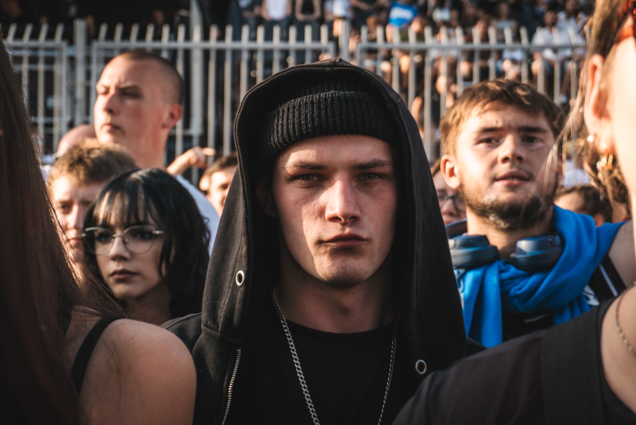 Portret młodego mężczyzny w czarnej bluzie z kapturem i czapce, otoczonego przez tłum ludzi na tle metalowej bariery.