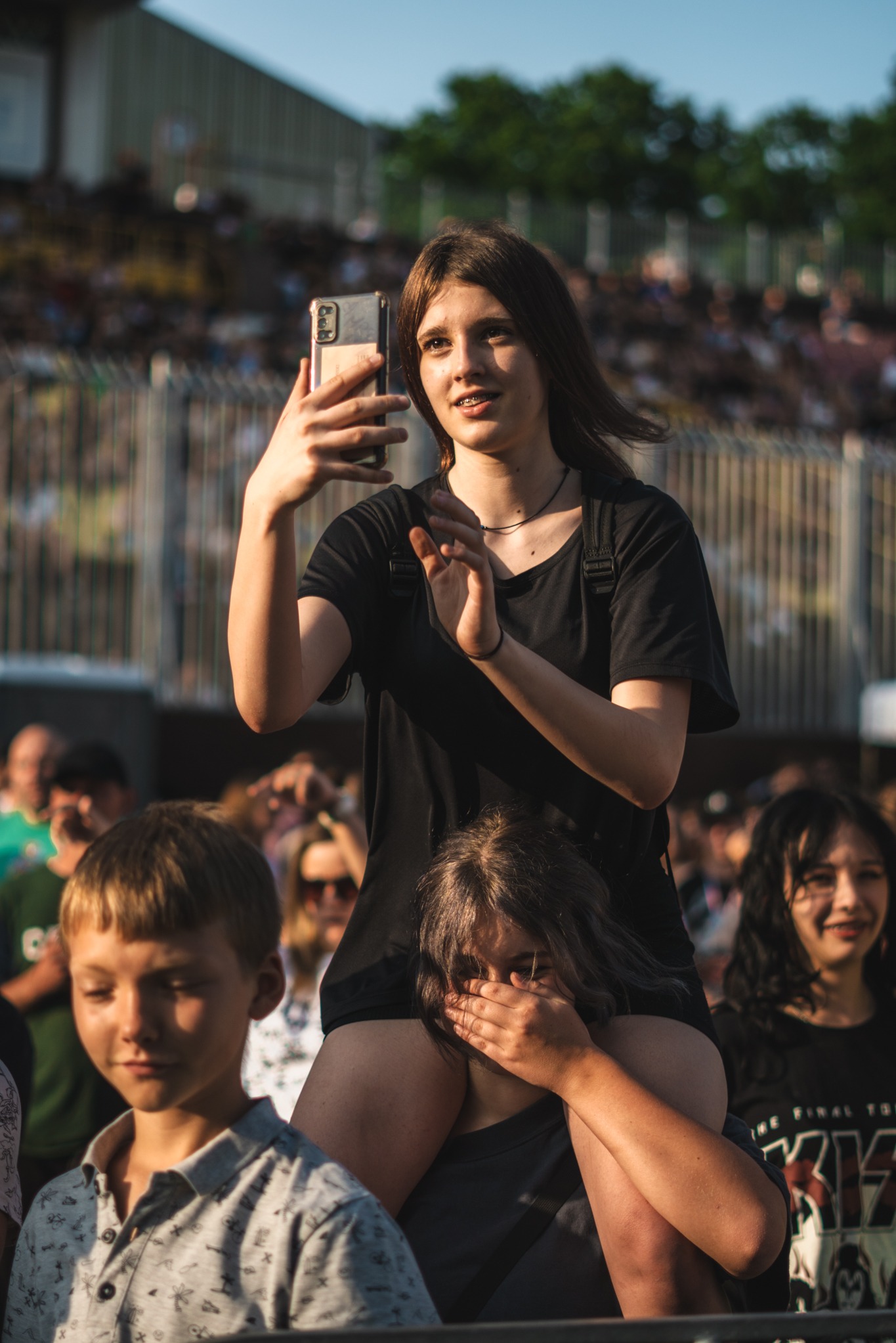 Dziewczyna na barana robi zdjęcie telefonem na tle rozmytego tłumu. Uśmiechnięta, z aparatem w dłoni, w czarnej koszulce i z aparatem na zębach.