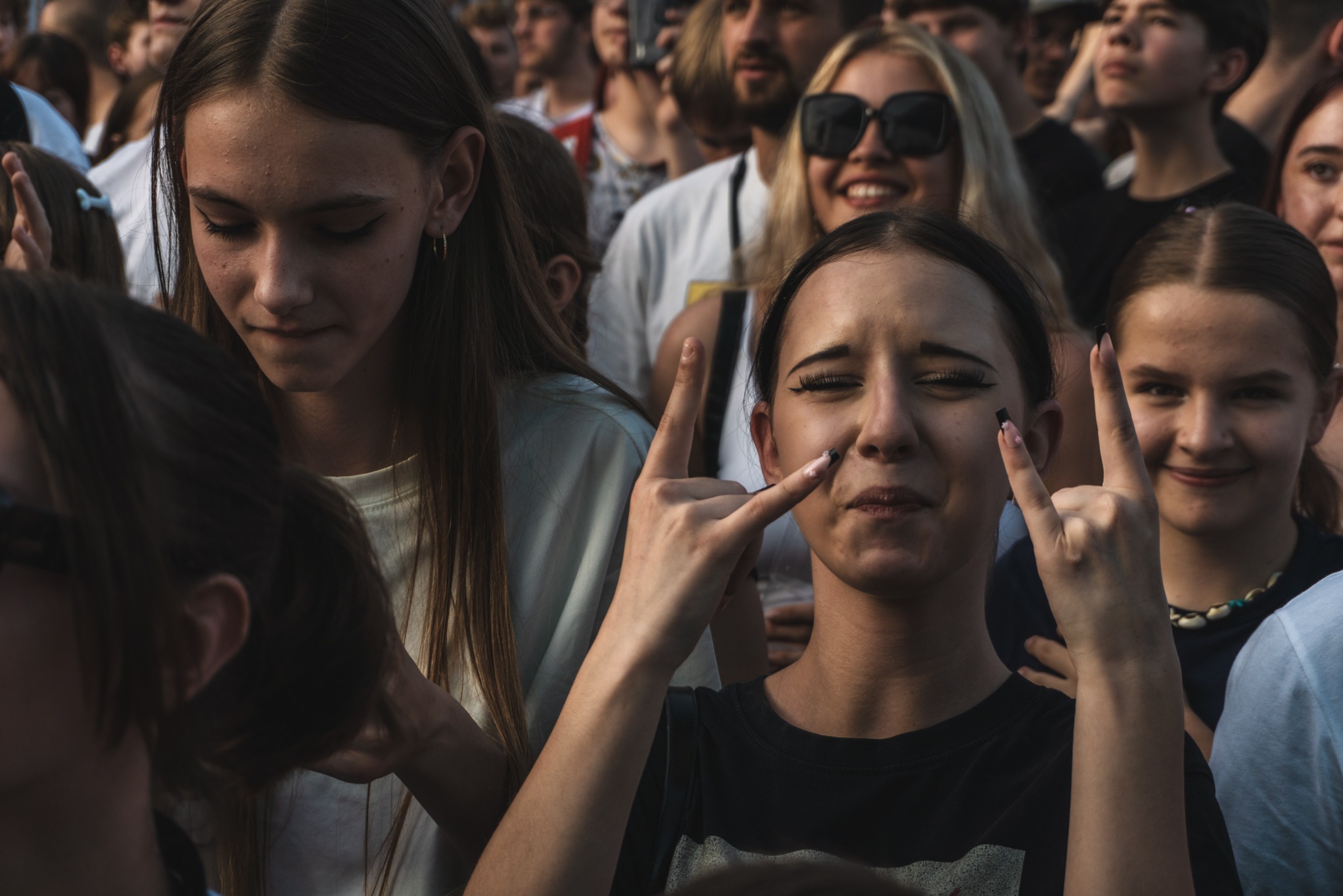 Tłum młodych ludzi, w centrum dziewczyna z makijażem i uniesionymi dłońmi, ukazująca gest rogów. Uśmiechnięte twarze i atmosfera koncertu.