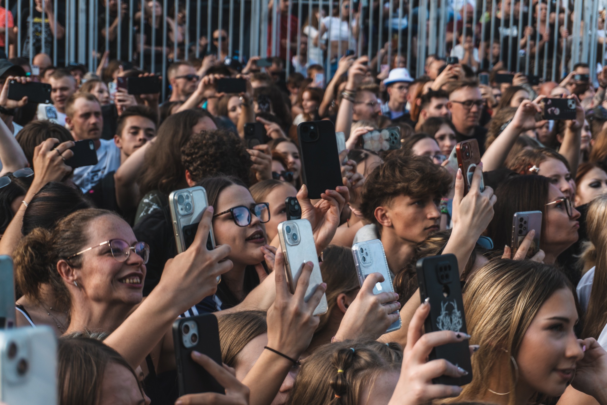 Tłum ludzi na koncercie, wielu z nich trzyma telefony komórkowe, nagrywając lub robiąc zdjęcia sceny, widoczne barierki w tle, różnorodne etui na telefonach.