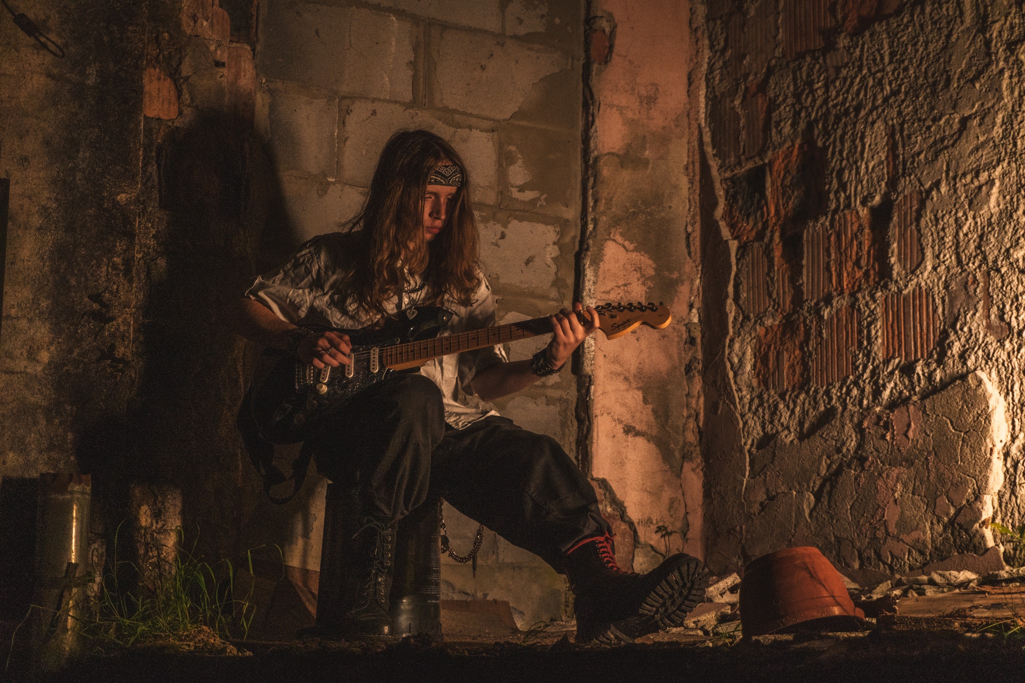 Młody muzyk z długimi włosami gra na gitarze elektrycznej w surowym wnętrzu z ceglanymi ścianami, oświetlonym ciepłym światłem. Wyrazisty styl i artystyczny klimat.