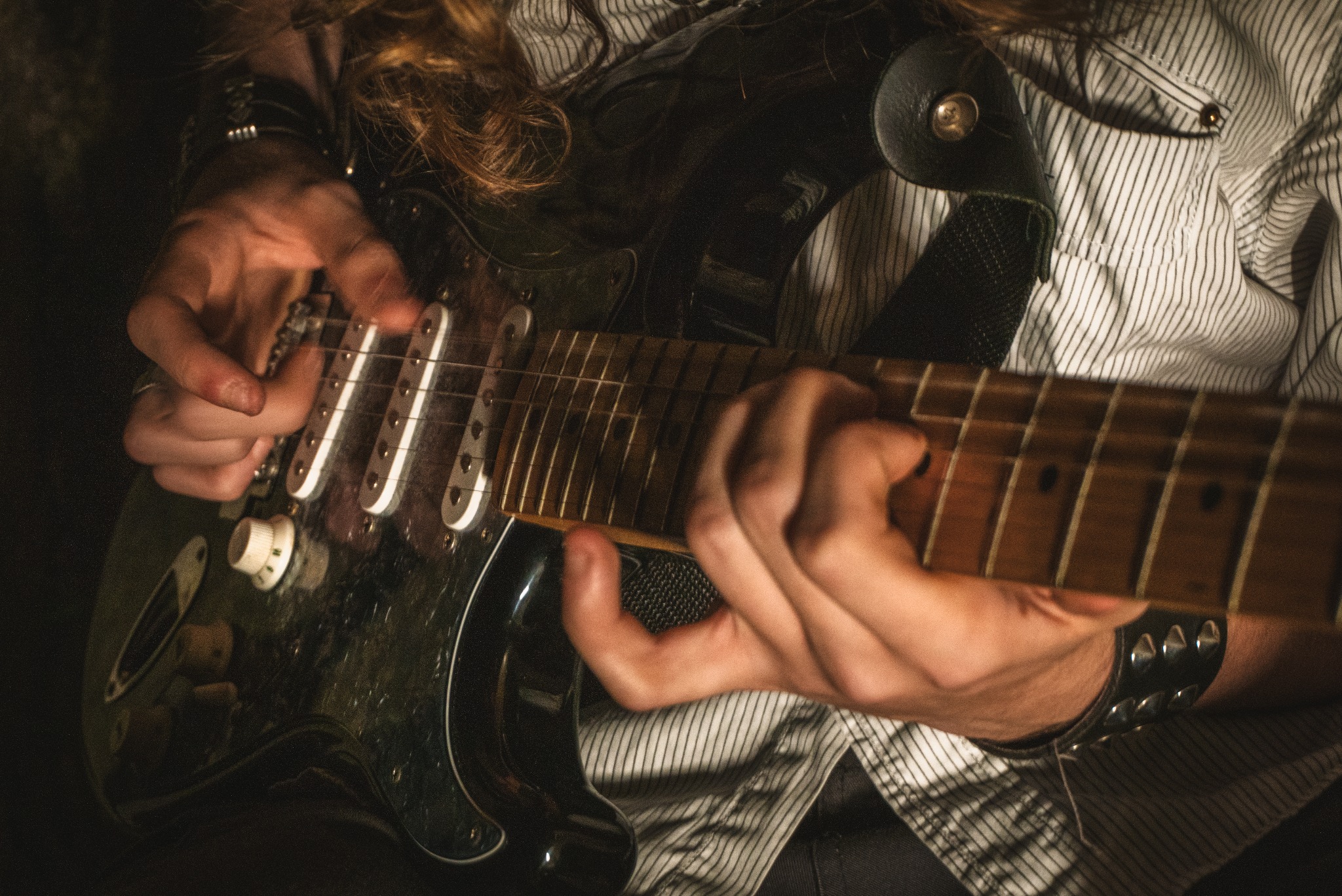 Zbliżenie na ręce gitarzysty grającego na elektrycznej gitarze typu Stratocaster, z widocznym paskiem i koszulą w paski. Dynamiczne ujęcie, ciemne tło.