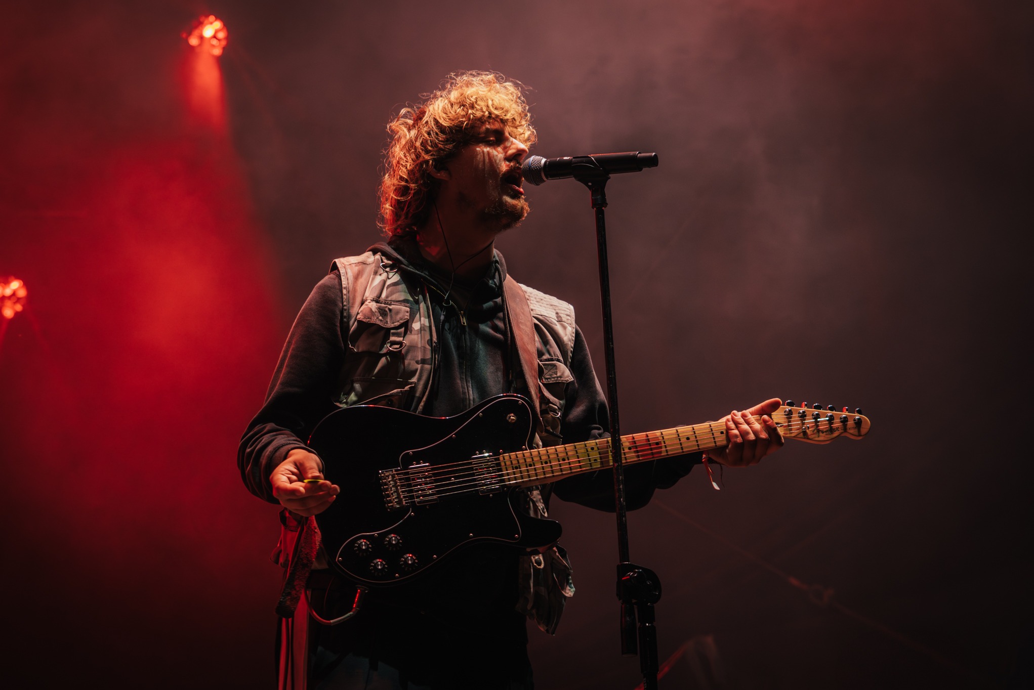 Mężczyzna z kręconymi włosami śpiewa do mikrofonu, grając na czarnej gitarze elektrycznej w czerwonym oświetleniu scenicznym podczas koncertu.