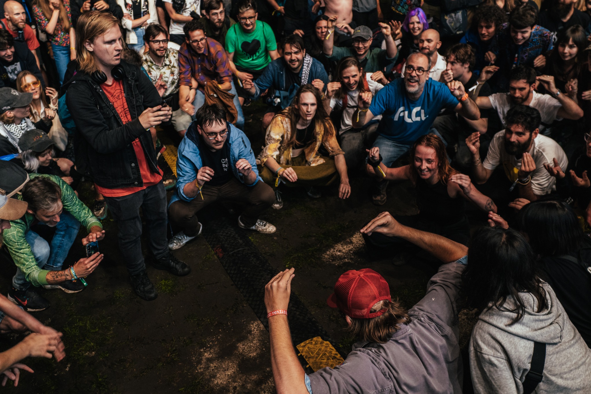 Zdjęcie z góry przedstawia grupę ludzi na festiwalu, niektórzy siedzą w kucki, inni stoją, z uniesionymi rękami, w centrum błotniste podłoże. W tle tłum.