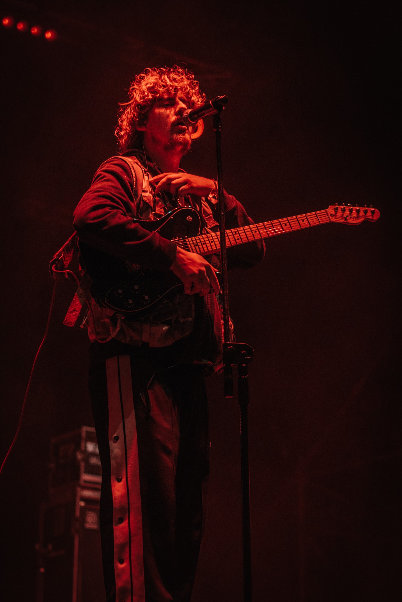 Wokalista z gitarą na scenie w czerwonym oświetleniu podczas występu na żywo. Widoczny mikrofon i fragmenty sprzętu muzycznego. Ujęcie z dołu.