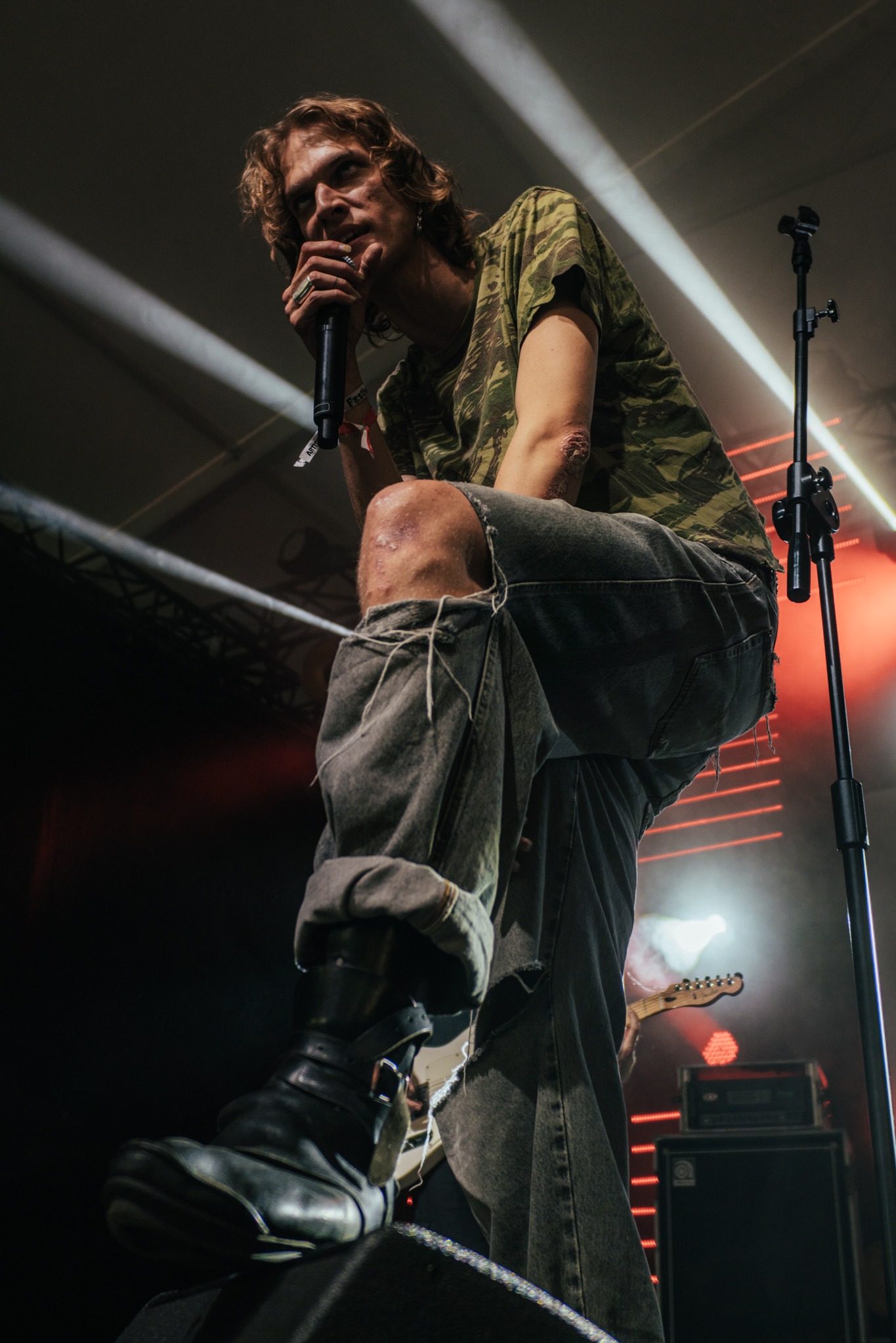 Wokalista z mikrofonem na scenie podczas koncertu, ujęcie z dołu, widoczne oświetlenie sceniczne i fragment gitary. Artysta w podartych jeansach i koszulce moro.