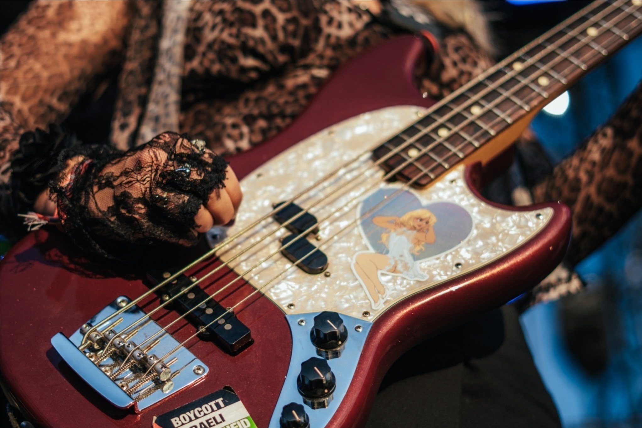 Kobieta w koronkowej rękawiczce gra na czerwonej gitarze basowej z naklejką pin-up girl. Ubrana w bluzkę w panterkę. Widoczne struny, przetworniki i naklejki na gitarze.