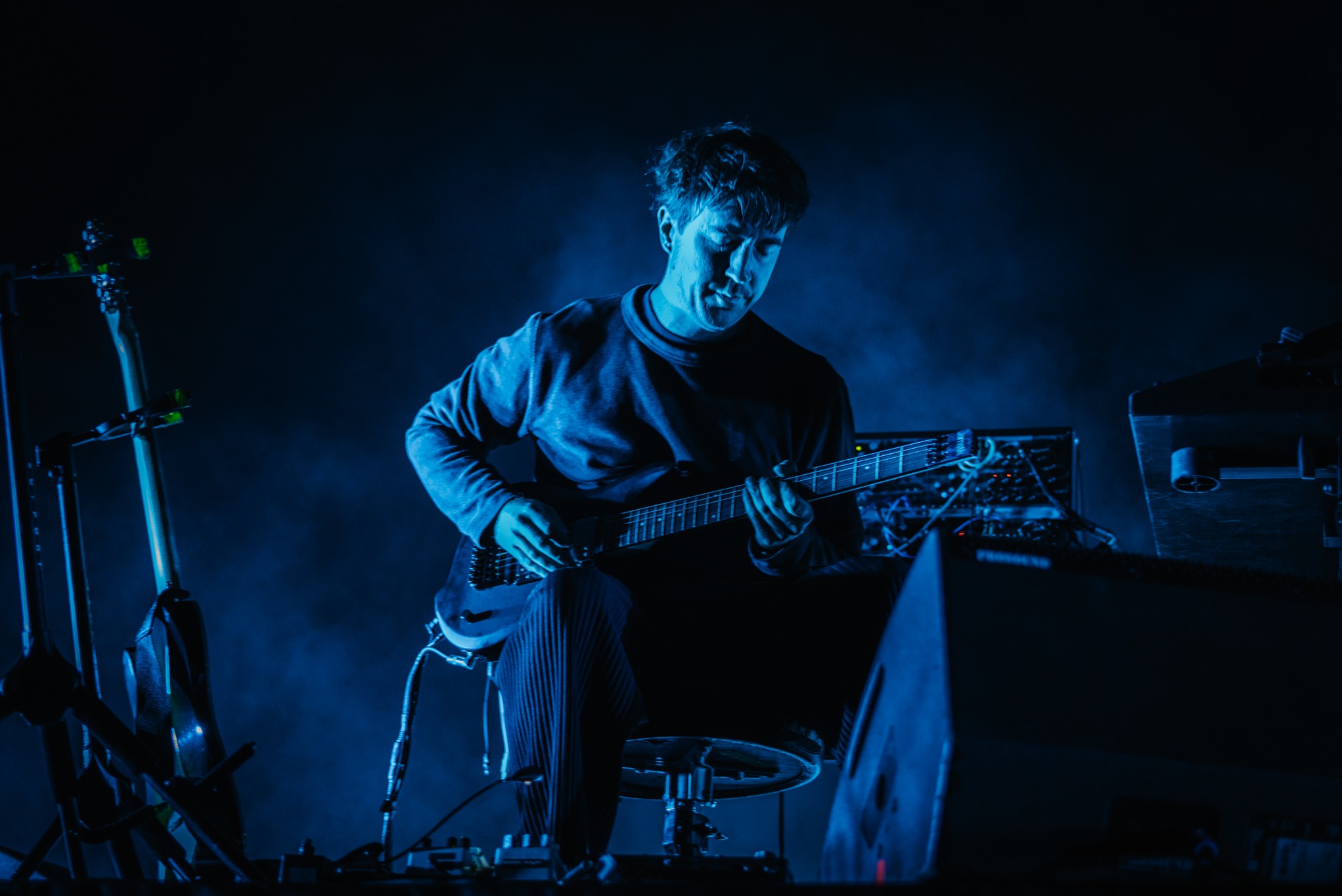 Muzyk grający na gitarze elektrycznej podczas koncertu, oświetlony intensywnym, niebieskim światłem. Widoczne instrumenty i sprzęt muzyczny na scenie.