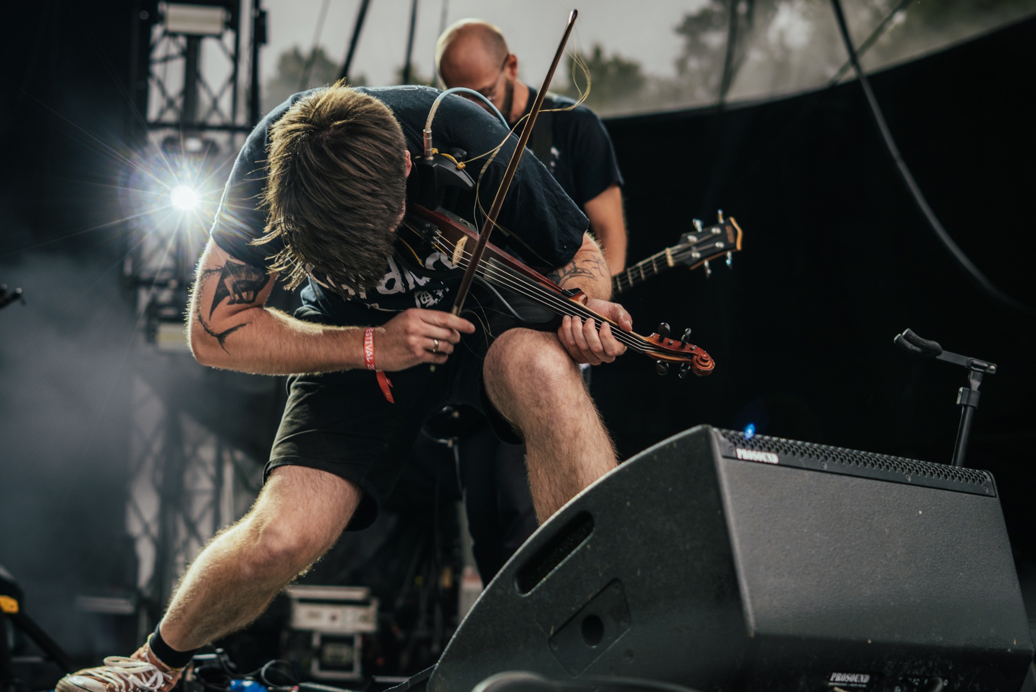 Skoncentrowany muzyk z tatuażami gra na skrzypcach podczas występu na żywo na scenie festiwalowej, z gitarzystą w tle i sprzętem nagłośnieniowym na pierwszym planie.