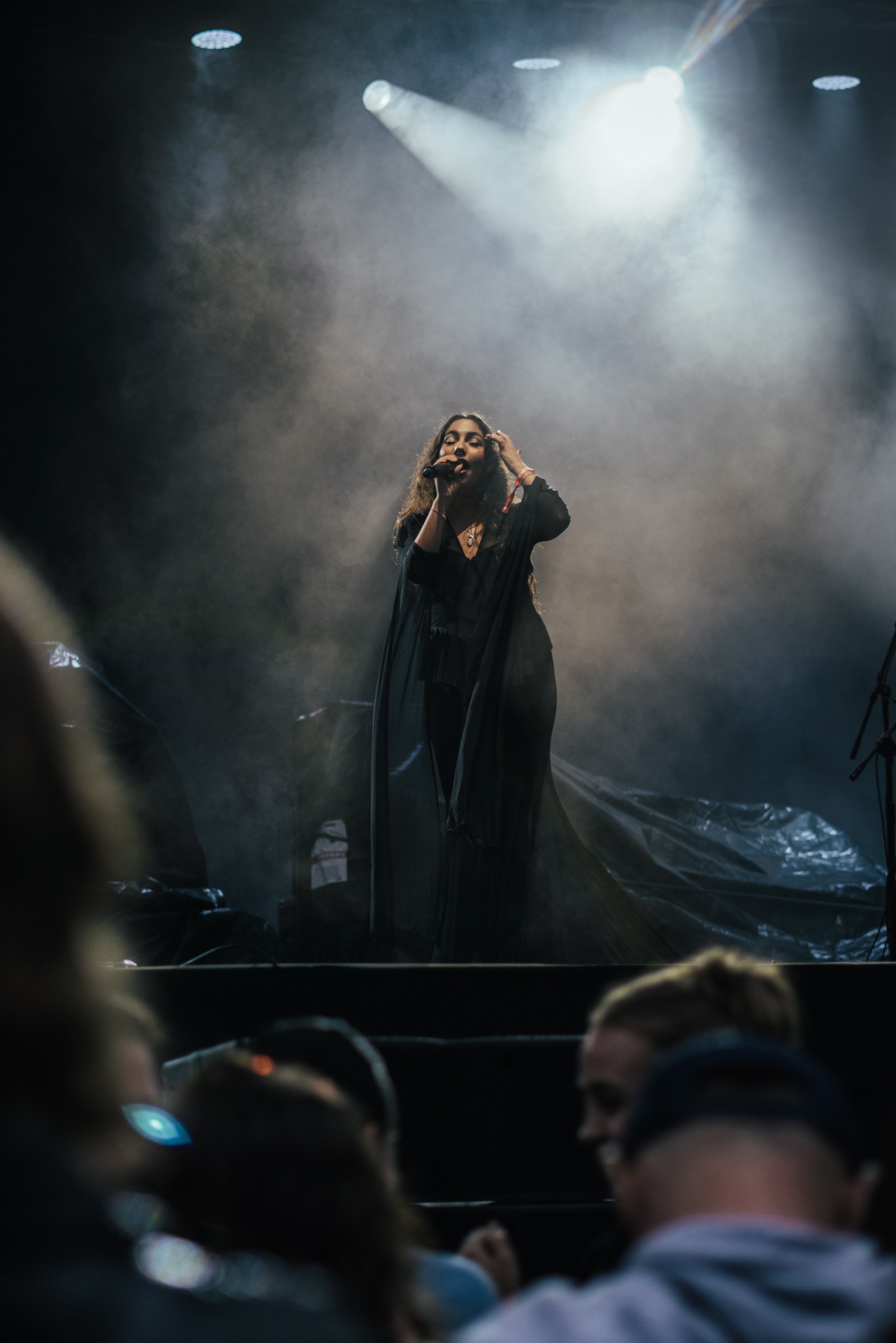 Wokalistka z mikrofonem na scenie w dymie i świetle reflektorów, widownia w tle. Ujęcie z dołu, dynamiczna atmosfera koncertu.
