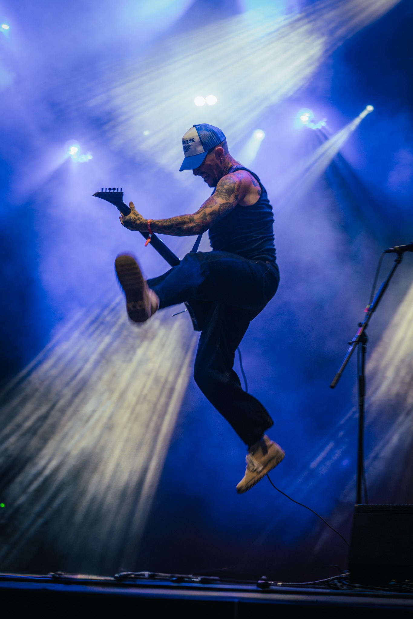Skaczący gitarzysta z tatuażami na scenie w niebieskim oświetleniu podczas koncertu. Dynamiczne ujęcie z podskoku z uniesioną nogą i gitarą.