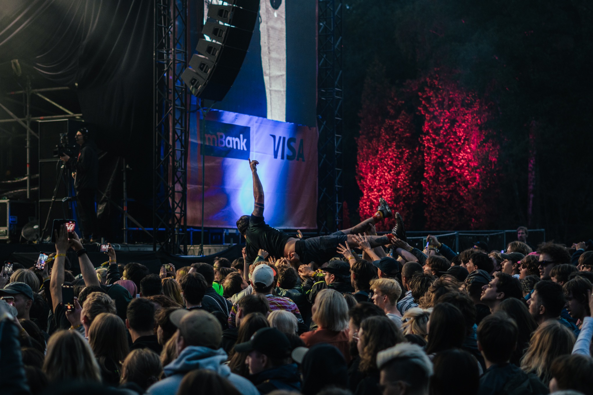 Tłum ludzi na koncercie, unoszący osobę nad głowami. W tle scena z ekranem i logotypami sponsorów, oświetlona czerwonymi światłami. Widoczne telefony nagrywające wydarzenie.