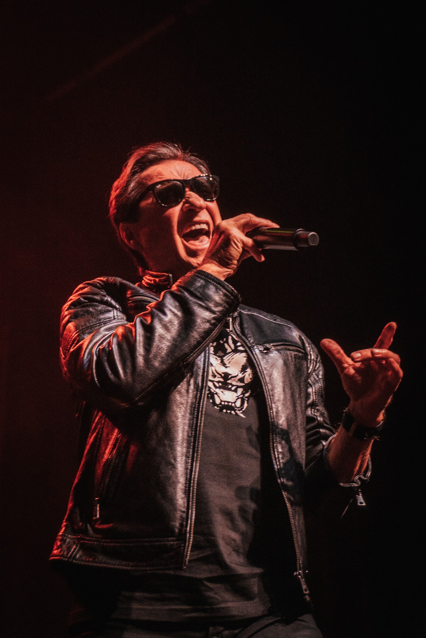 Wokalista w skórzanej kurtce, śpiewa do mikrofonu na scenie, oświetlony czerwonym światłem. Widoczne okulary przeciwsłoneczne i ekspresja na twarzy.