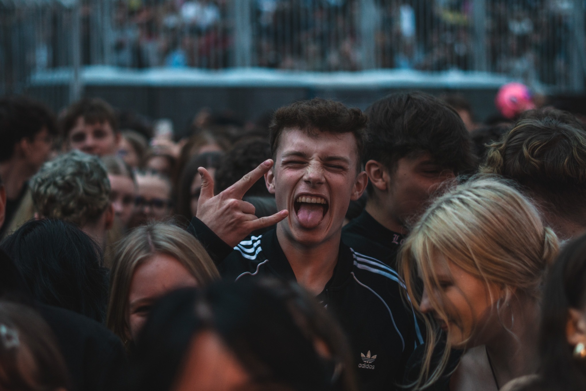 Rozentuzjazmowany młody mężczyzna w tłumie, pokazuje gest dłońmi, wykrzywia twarz i wystawia język. Widoczny tłum ludzi w tle. Zdjęcie z imprezy masowej.