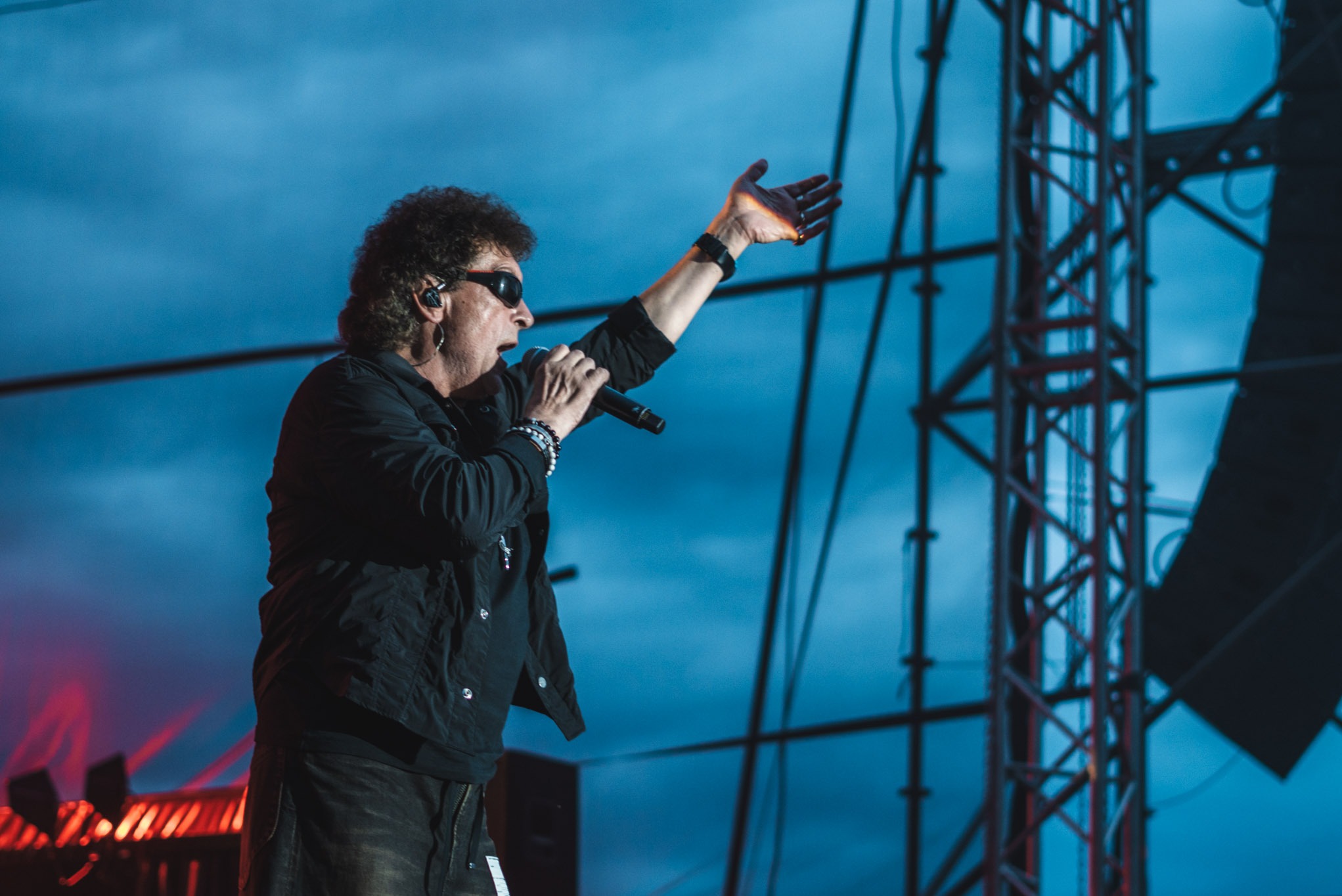 Wokalista z mikrofonem na scenie podczas koncertu plenerowego, w tle konstrukcja sceniczna i niebieskie oświetlenie. Ujęcie z dołu, dynamiczne.