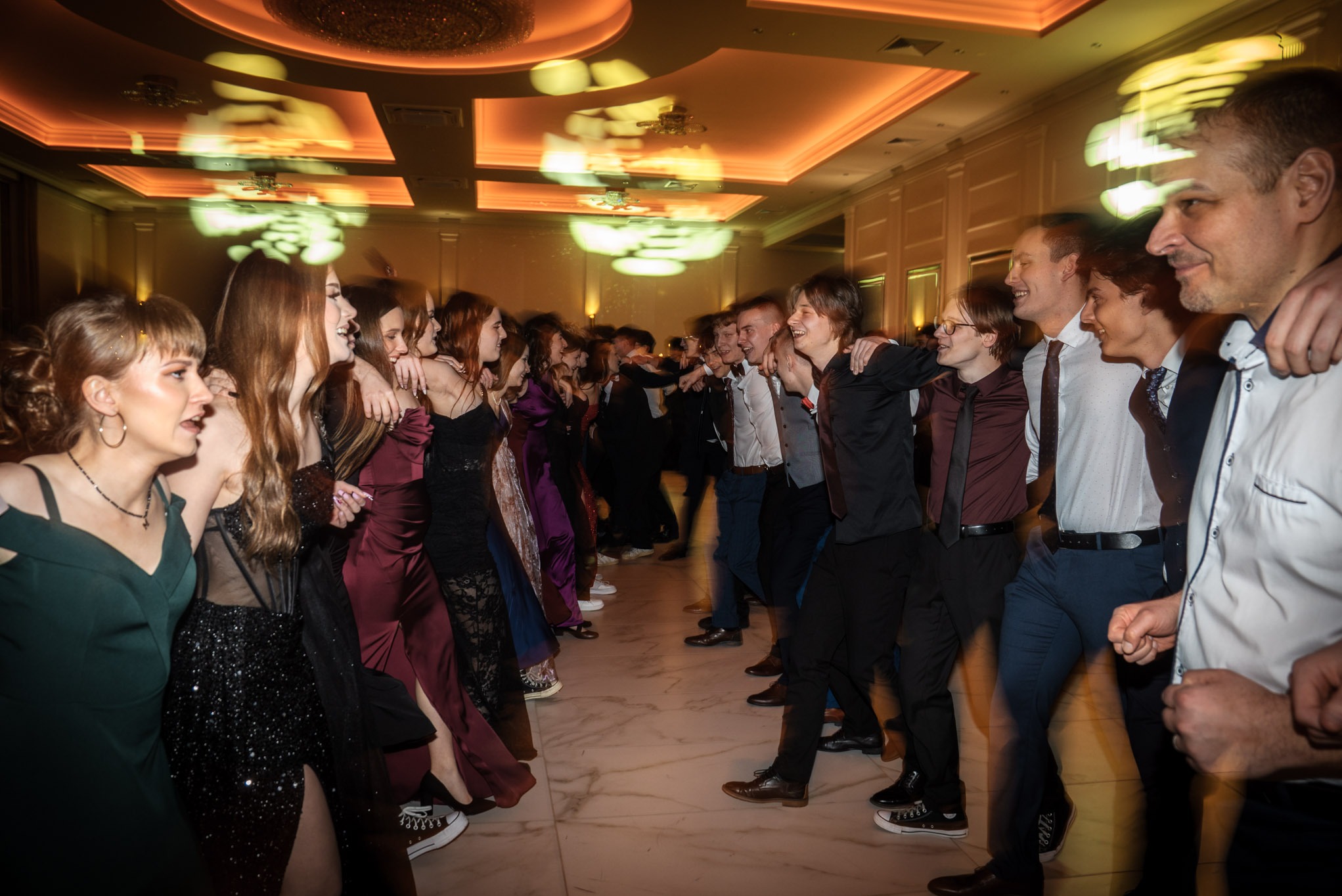 Radosny korowód maturzystów w eleganckich strojach, tańczących w sali balowej z marmurową podłogą i dekoracyjnym oświetleniem podczas studniówki.