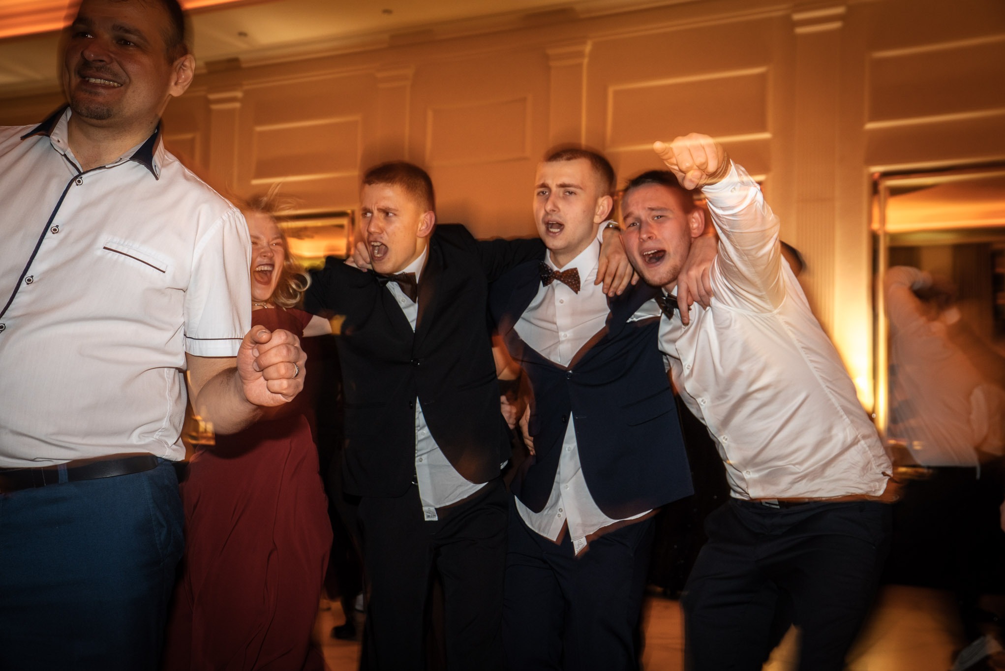 Rozmazane ujęcie rozbawionych maturzystów w trakcie studniówki, w ciemnych garniturach i z muszkami, w dynamicznych pozach, z uniesionymi rękami i otwartymi ustami.