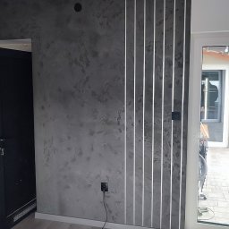 Perfect Home usługi ogólnobudowlane - Dekoracyjna ściana w odcieniach szarości z pionowymi, podświetlanymi listwami. Widoczny fragment podłogi z paneli i drzwi balkonowe.