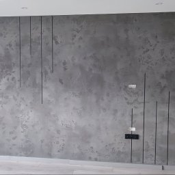 Perfect Home usługi ogólnobudowlane - Szara ściana z efektem dekoracyjnym i pionowymi czarnymi liniami. Widoczne gniazdka elektryczne i listwa przypodłogowa. Nowoczesny design wnętrza.