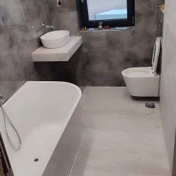 Perfect Home usługi ogólnobudowlane - Nowoczesna łazienka z wanną, umywalką na blacie i podwieszaną toaletą. Szare płytki imitujące marmur na ścianach, jasna podłoga. Okno z widokiem na zewnątrz.