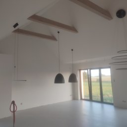 Perfect Home usługi ogólnobudowlane - Jasne wnętrze z wysokim stropem i drewnianymi belkami. Widoczne nowoczesne lampy wiszące, duże okna i świeżo pomalowane ściany. Na podłodze widoczne ślady po pracach remontowych.