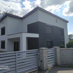 Perfect Home usługi ogólnobudowlane - Dwukondygnacyjny dom z szarą elewacją, roletami zewnętrznymi i jasnym ogrodzeniem panelowym. Widoczne fragmenty zieleni i błękitne niebo z chmurami.