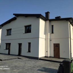 Perfect Home usługi ogólnobudowlane - Dwukondygnacyjny dom z jasną elewacją i ciemną stolarką okienną, widoczny z poziomu brukowanego podjazdu w słoneczny dzień. Widoczne detale wykończenia.