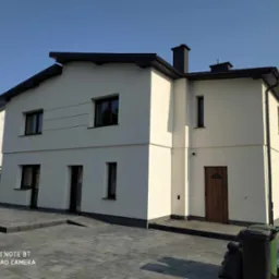 Dwukondygnacyjny dom z jasną elewacją i ciemną stolarką okienną, widoczny z poziomu brukowanego podjazdu w słoneczny dzień. Widoczne detale wykończenia.