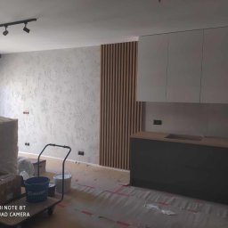 Perfect Home usługi ogólnobudowlane - Nowoczesna kuchnia w trakcie wykończenia: szare szafki, drewniane lamele na ścianie, dekoracyjna faktura ściany. Podłoga zabezpieczona folią malarską.