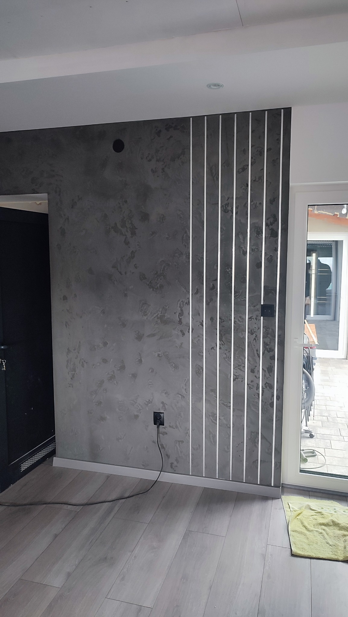 Dekoracyjna ściana w odcieniach szarości z pionowymi, podświetlanymi listwami. Widoczny fragment podłogi z paneli i drzwi balkonowe.