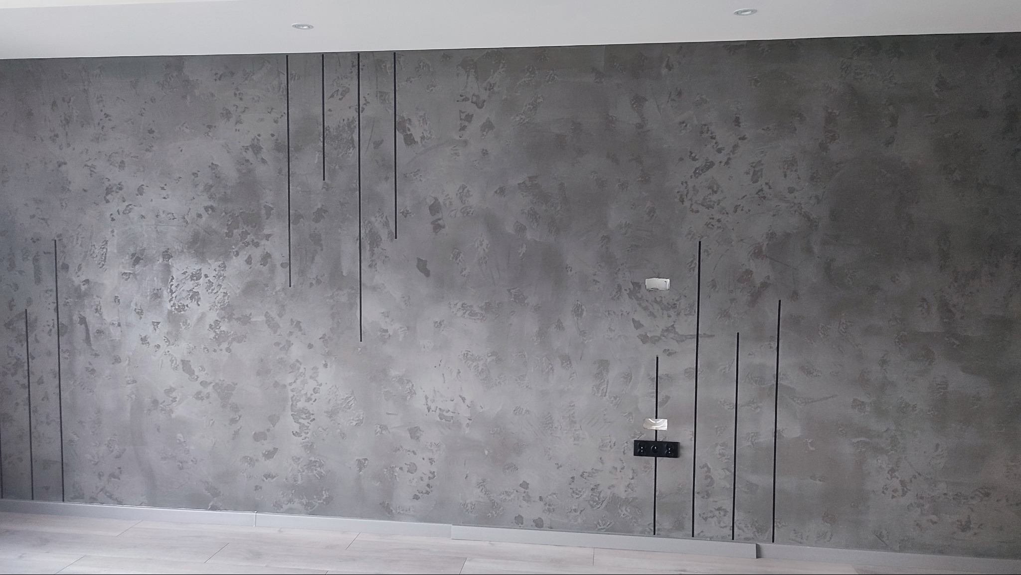 Szara ściana z efektem dekoracyjnym i pionowymi czarnymi liniami. Widoczne gniazdka elektryczne i listwa przypodłogowa. Nowoczesny design wnętrza.