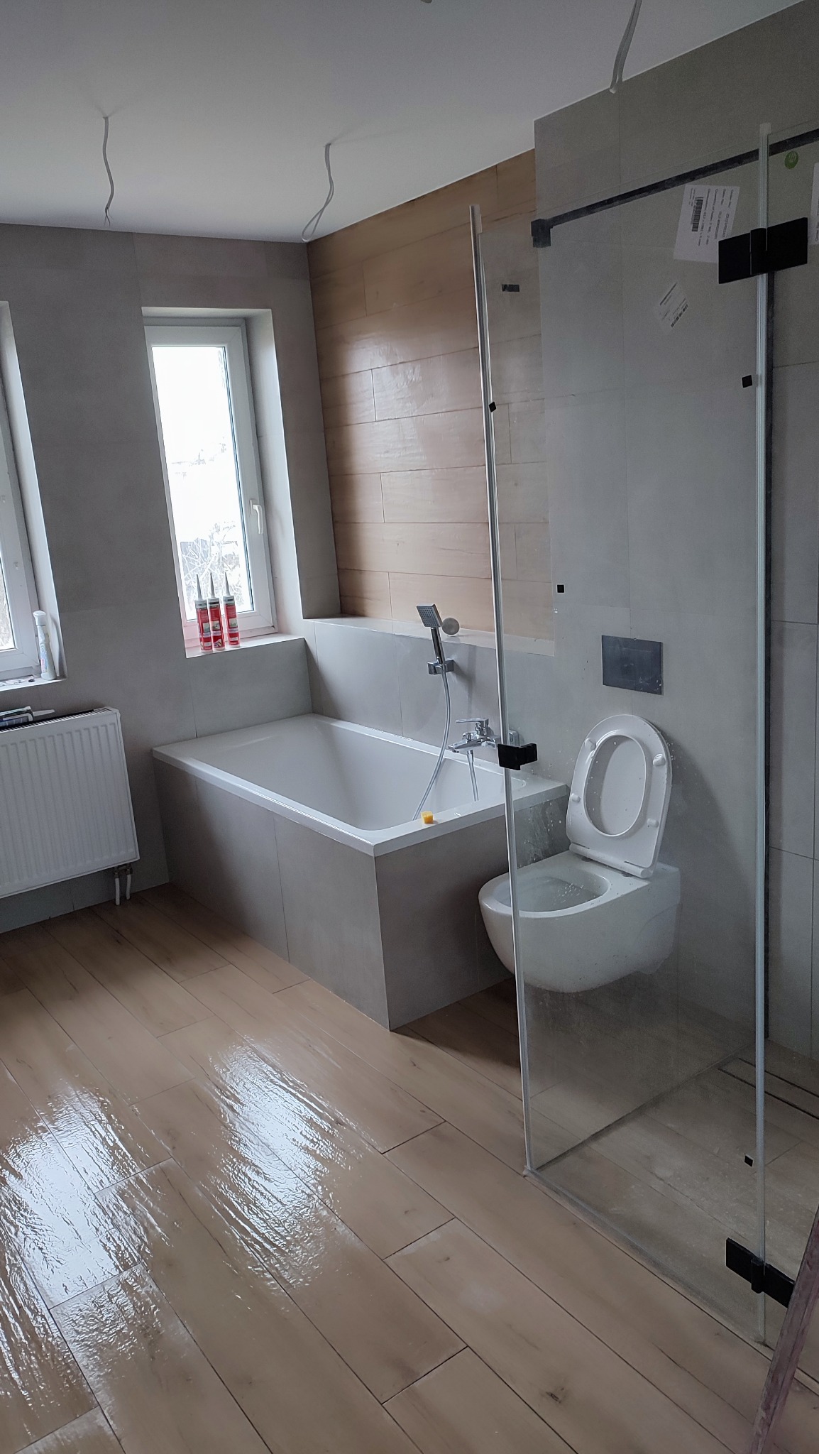 Wykończona łazienka z wanną w zabudowie, toaletą i szklanym panelem prysznicowym. Widoczne drewnopodobne płytki na podłodze i częściowo na ścianie, okno z kosmetykami.