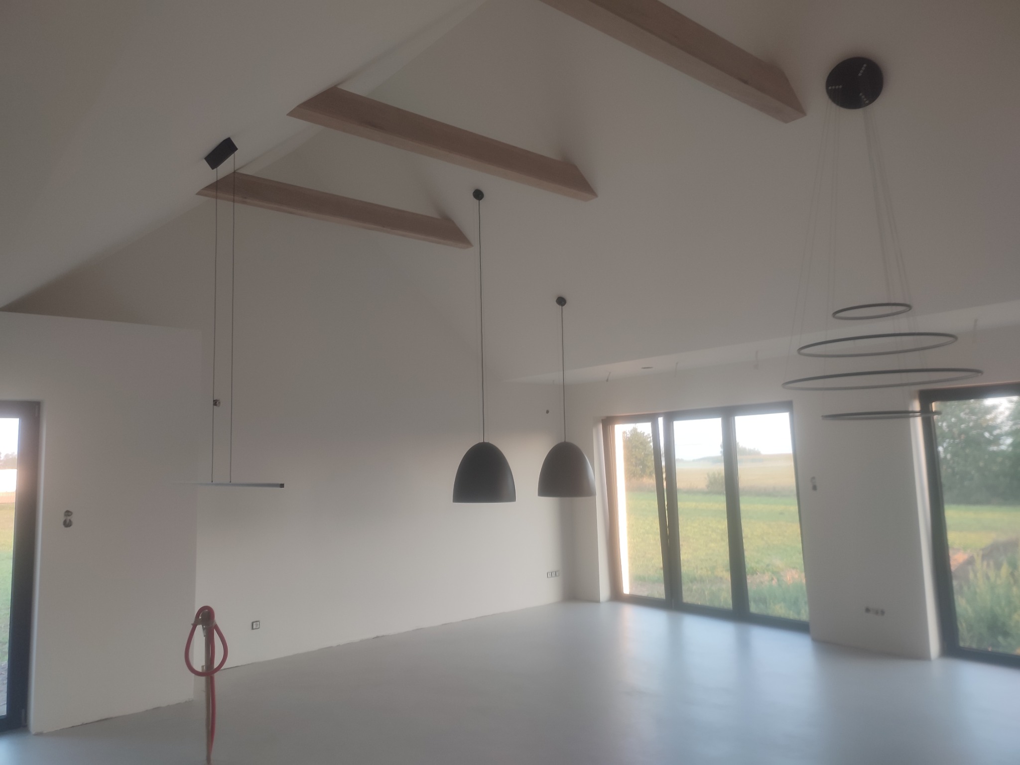 Jasne wnętrze z wysokim stropem i drewnianymi belkami. Widoczne nowoczesne lampy wiszące, duże okna i świeżo pomalowane ściany. Na podłodze widoczne ślady po pracach remontowych.