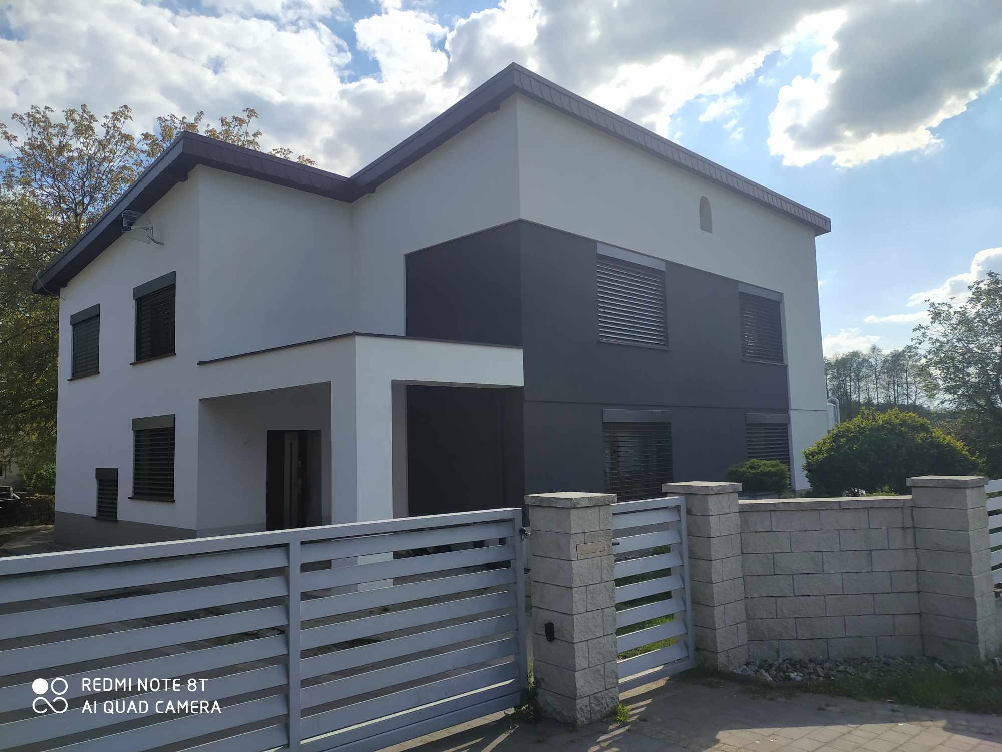 Dwukondygnacyjny dom z szarą elewacją, roletami zewnętrznymi i jasnym ogrodzeniem panelowym. Widoczne fragmenty zieleni i błękitne niebo z chmurami.