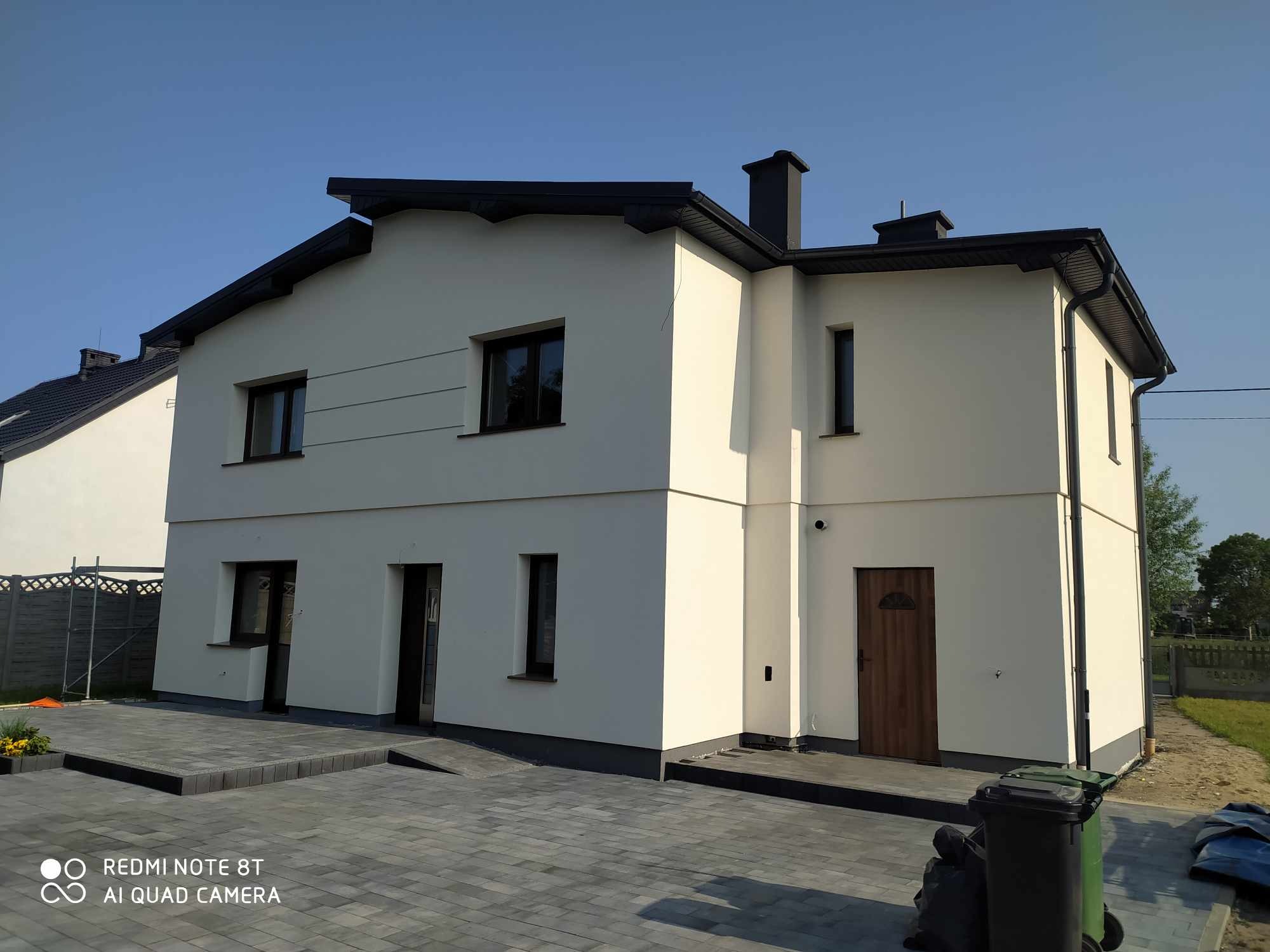 Dwukondygnacyjny dom z jasną elewacją i ciemną stolarką okienną, widoczny z poziomu brukowanego podjazdu w słoneczny dzień. Widoczne detale wykończenia.