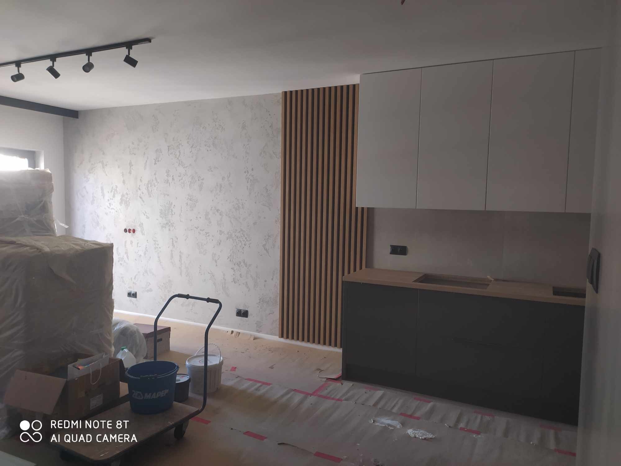 Nowoczesna kuchnia w trakcie wykończenia: szare szafki, drewniane lamele na ścianie, dekoracyjna faktura ściany. Podłoga zabezpieczona folią malarską.