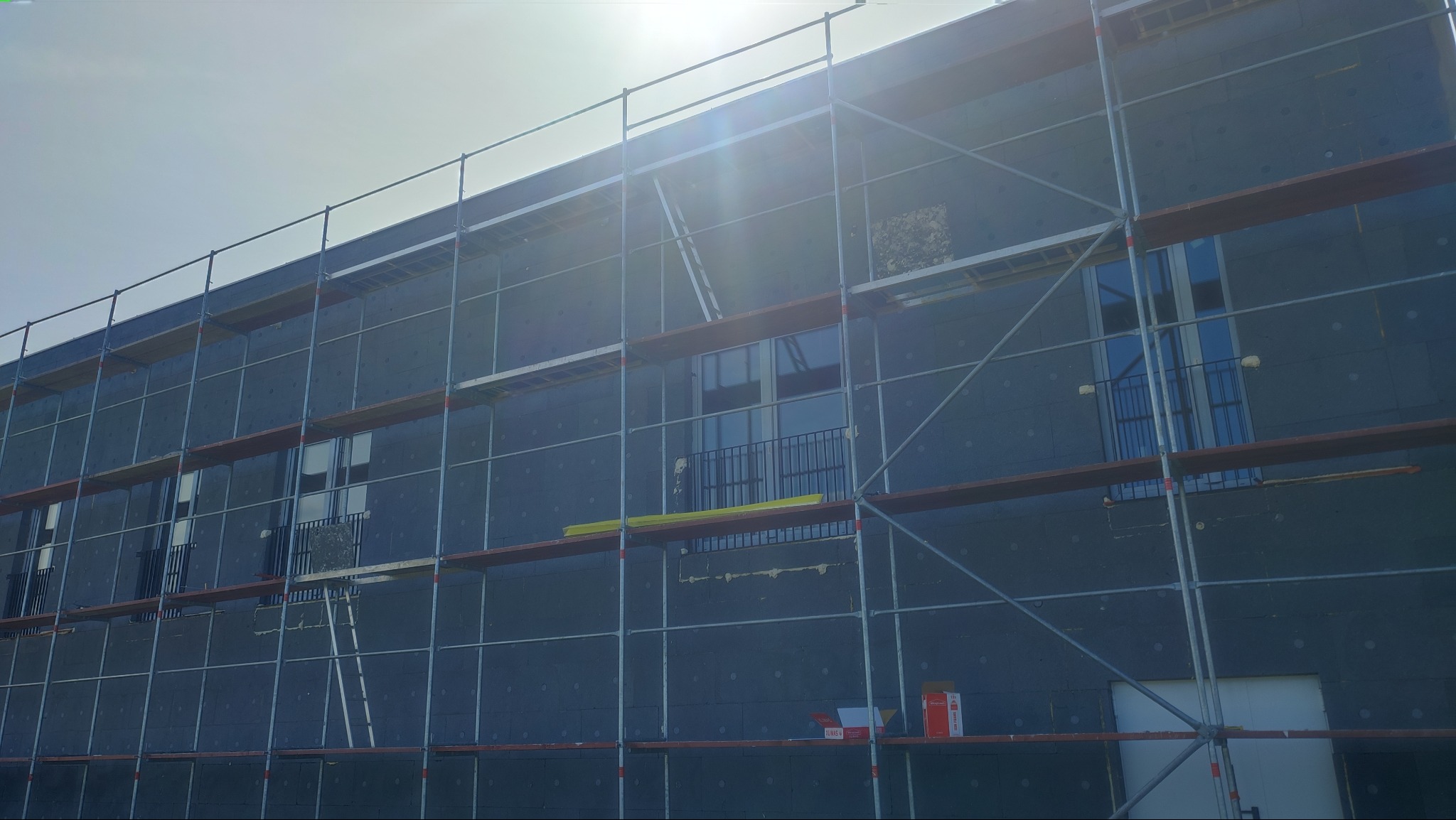 Elewacja budynku w trakcie ocieplania, widoczne rusztowania, okna z barierkami i materiały budowlane. Prace na wysokościach w słoneczny dzień.