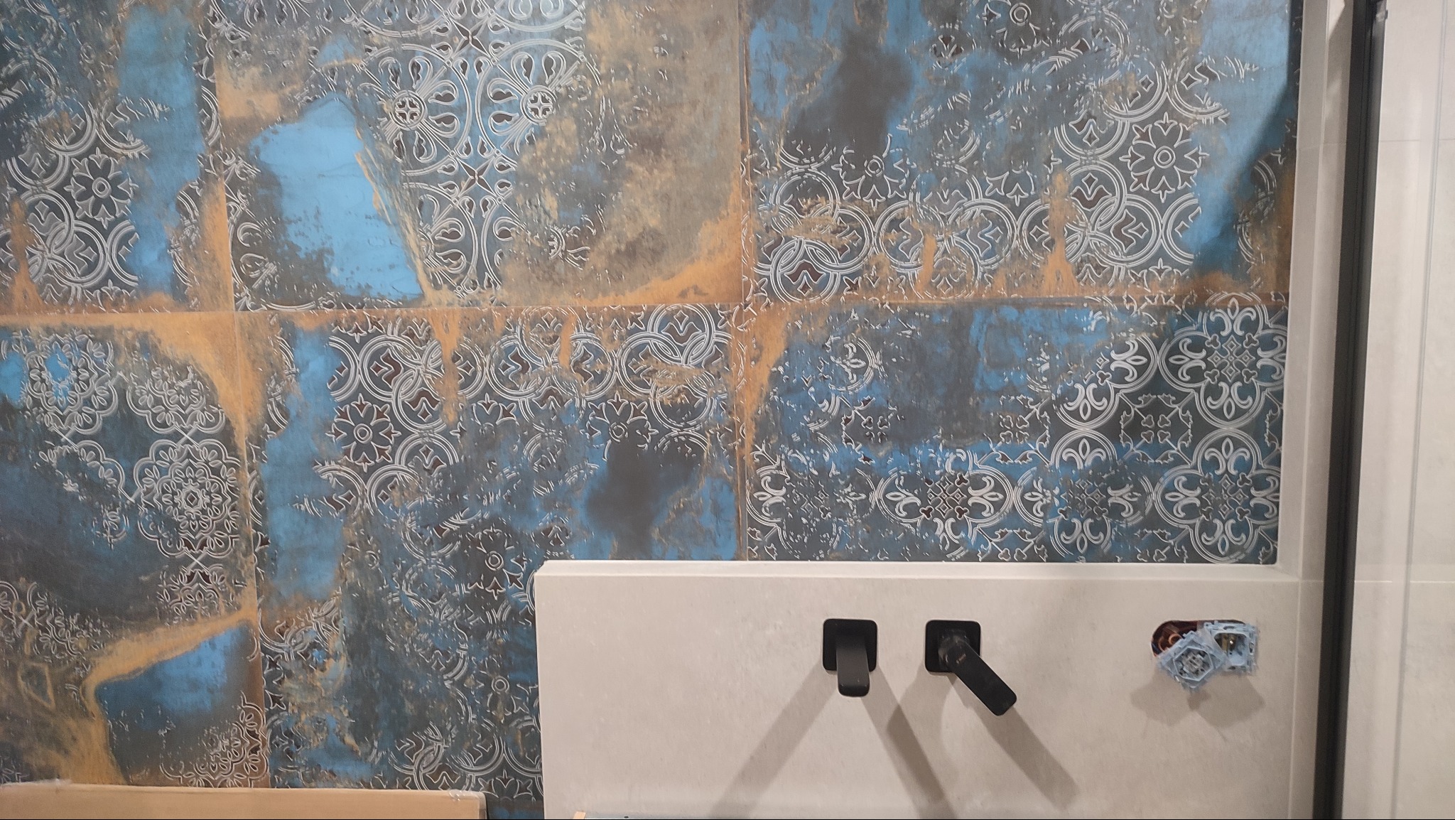 Wykończona ściana w łazience z dekoracyjnymi płytkami w odcieniach błękitu i złota, bateria umywalkowa w kolorze białym z czarnymi elementami.