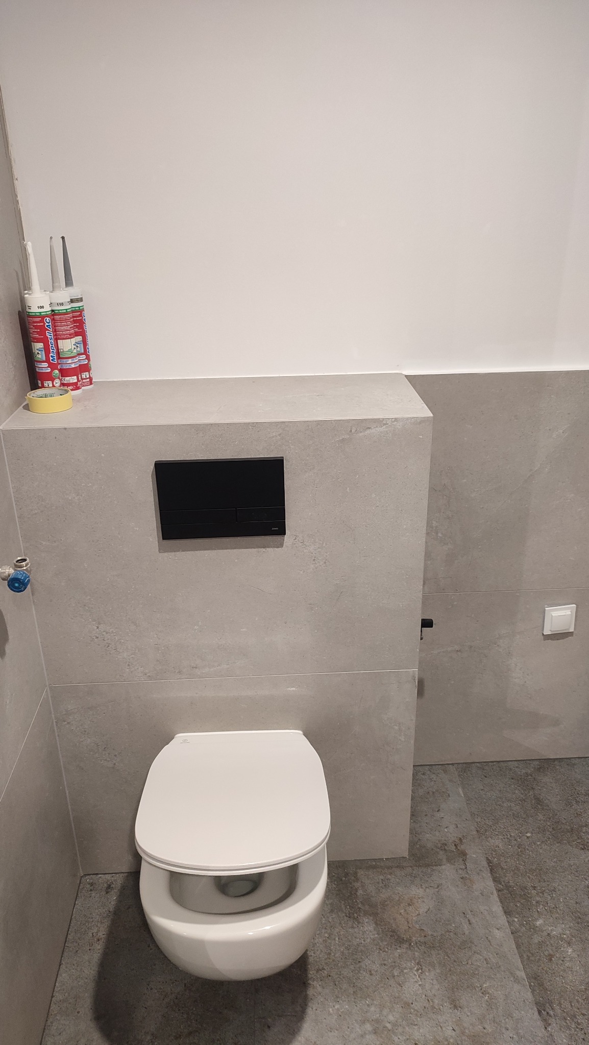 Nowoczesna toaleta podwieszana z czarnym panelem spłukującym, wykończona płytkami w odcieniach szarości. Widoczne silikony i taśma malarska.