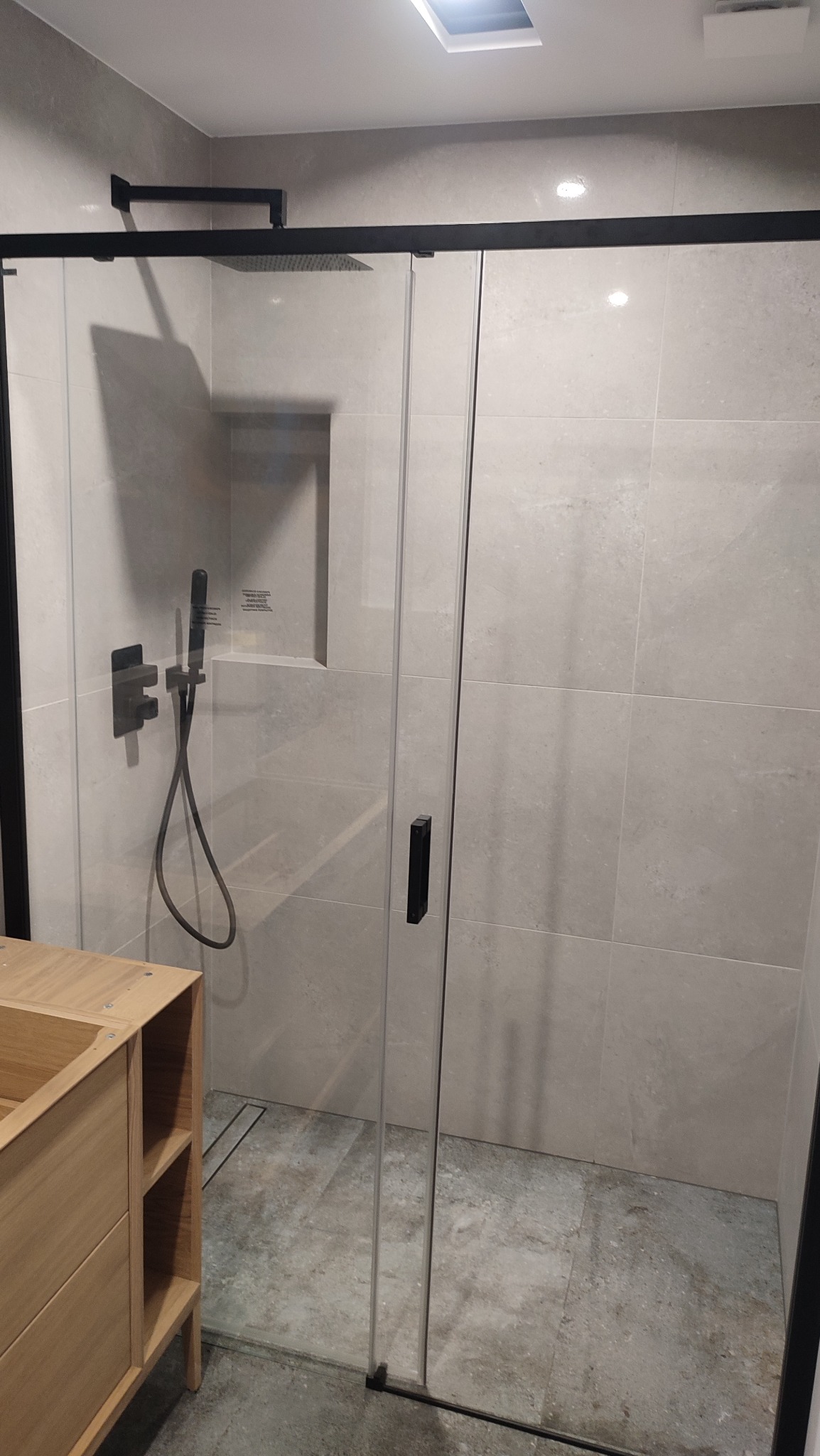 Nowoczesna kabina prysznicowa z czarnymi profilami i deszczownicą, szare płytki, drewniana szafka umywalkowa. Minimalistyczny design łazienki.