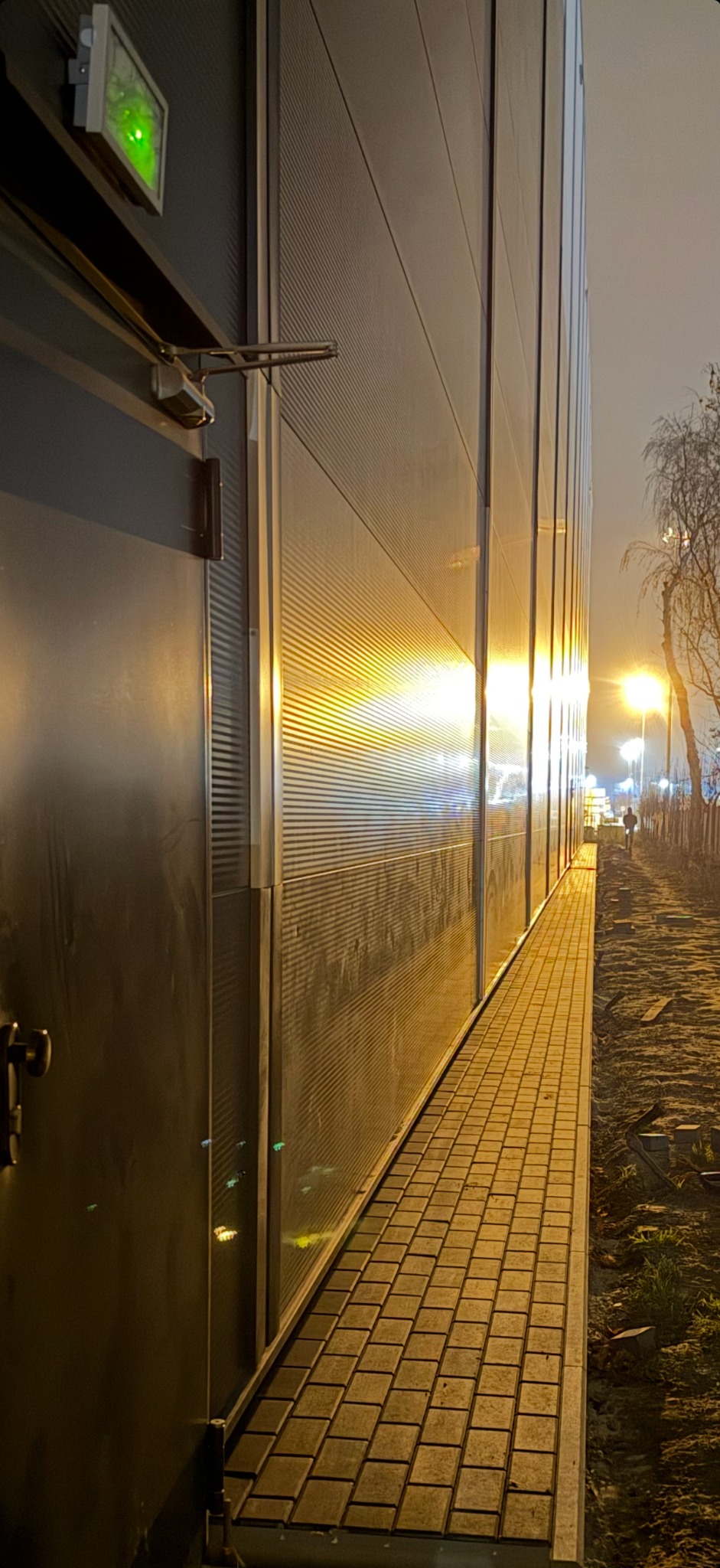 Oświetlona nocą ścieżka z kostki brukowej przylegająca do budynku z metalową fasadą. W tle widoczne latarnie i sylwetka osoby.